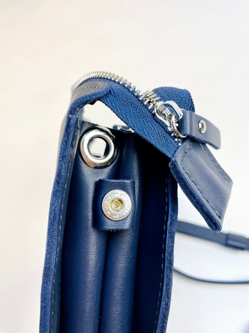 May include: A navy blue leather crossbody bag with a zipper closure and a silver metal ring. The bag has a flap with a silver metal button.