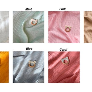K&ouml;nnte beinhalten: Eine Auswahl an Baby-Bei&szlig;ringen in den Farben Wei&szlig;, Mint, Rosa, Beige, Senf, Blau und Koralle. Jeder Bei&szlig;ring hat einen Holzring und einen b&auml;renf&ouml;rmigen Silikonaufsatz, der auf einem farblich passenden Stoffhintergrund liegt.