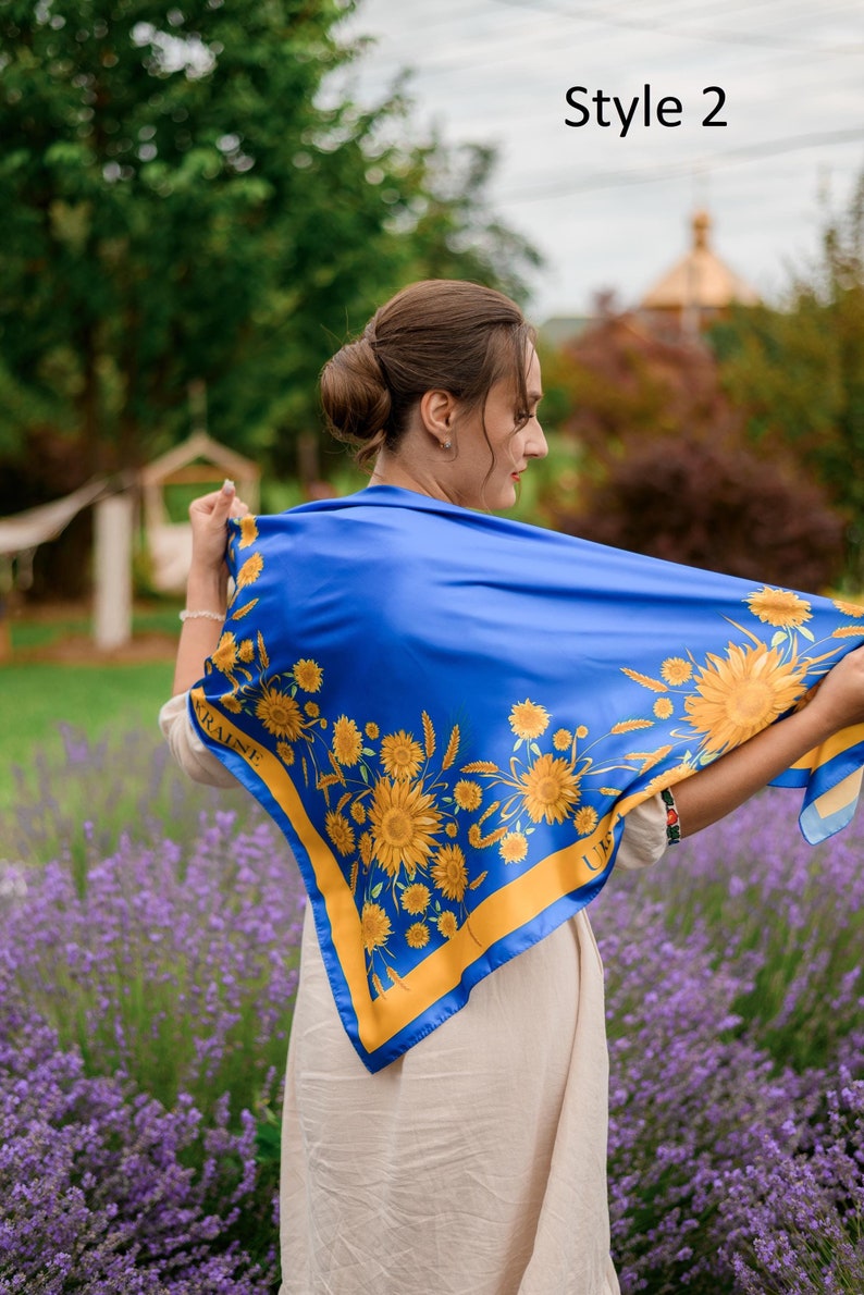 May include: A woman wearing a blue silk scarf with a yellow sunflower pattern and the word "UKRAINE" printed on it. The scarf is tied around her neck and she is standing in a field of lavender.