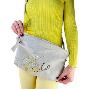May include: A light gray linen crossbody bag with an embroidered design of yellow dandelions and the name "Eva" in black thread.