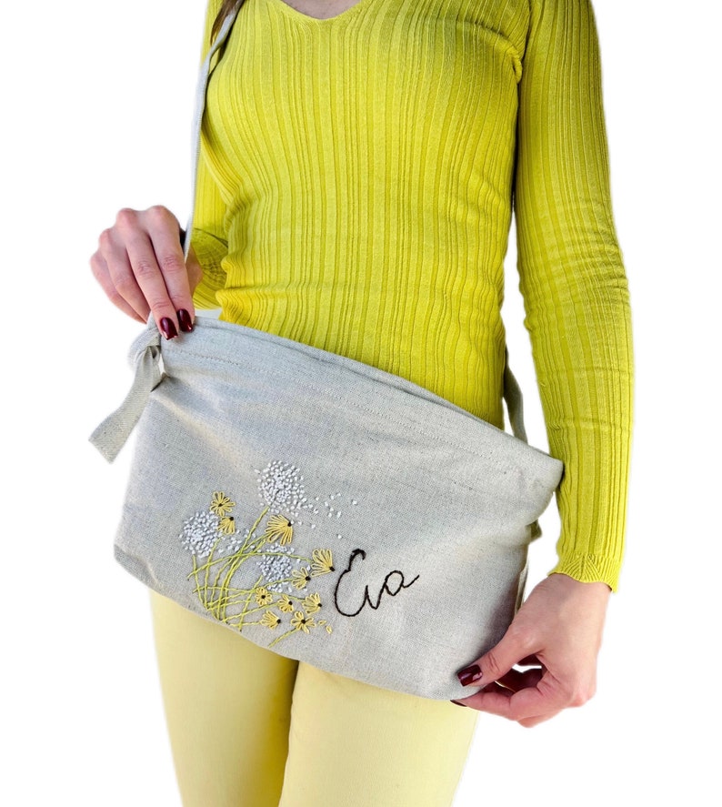May include: A light gray linen crossbody bag with an embroidered design of yellow dandelions and the name "Eva" in black thread.