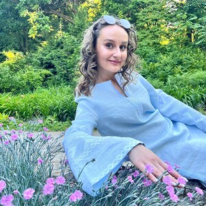 Op de afbeelding: Een vrouw in een lichtblauwe linnen jurk met witte borduursels zit in een tuin met roze bloemen.