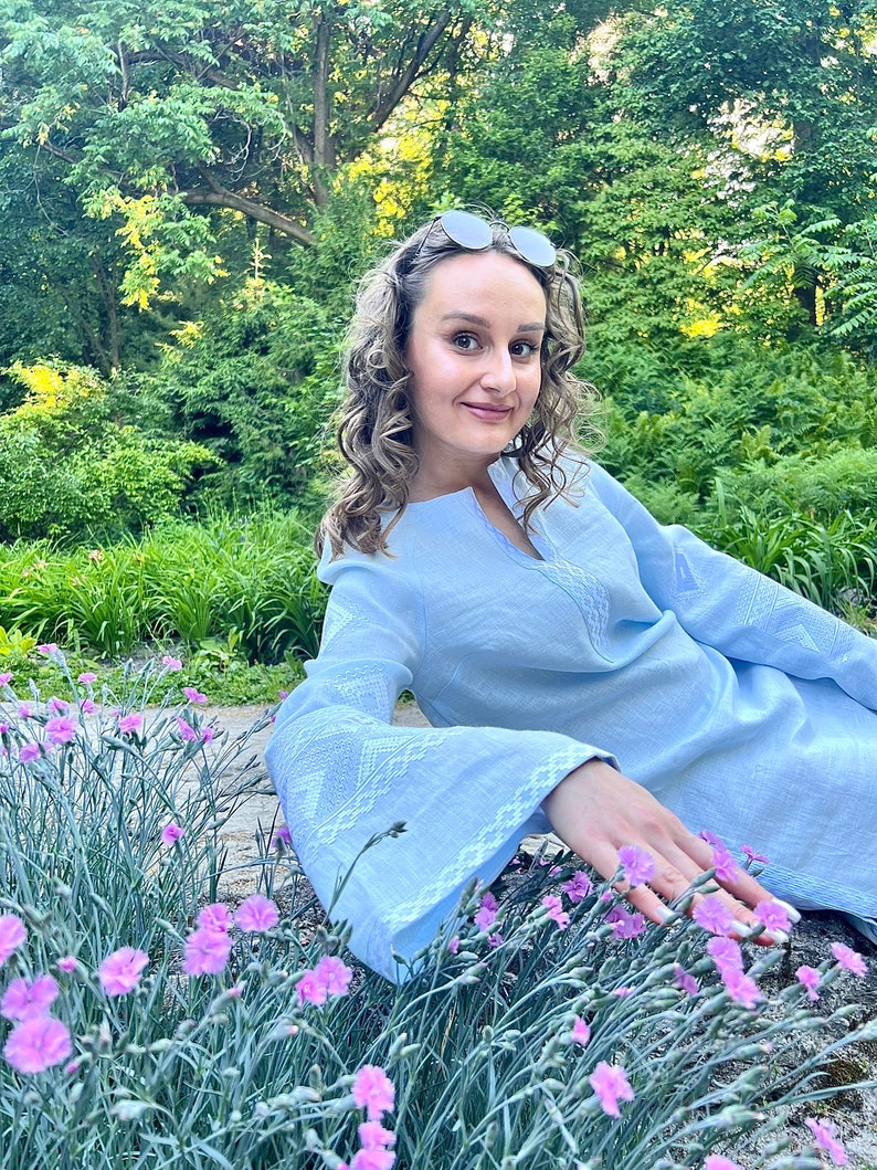 Op de afbeelding: Een vrouw in een lichtblauwe linnen jurk met witte borduursels zit in een tuin met roze bloemen.