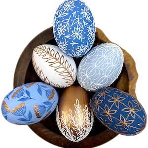 May include: A collection of decorative Easter eggs in a wooden bowl. The eggs are painted in various colors, including blue, white, and gold, and feature floral and leaf patterns. The eggs are approximately 3 inches tall.