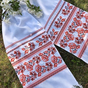 May include: Two white embroidered cloths with red floral patterns and fringed edges. The floral design includes roses and leaves. A sprig of white flowers rests on the cloth, which is displayed on a grassy surface.