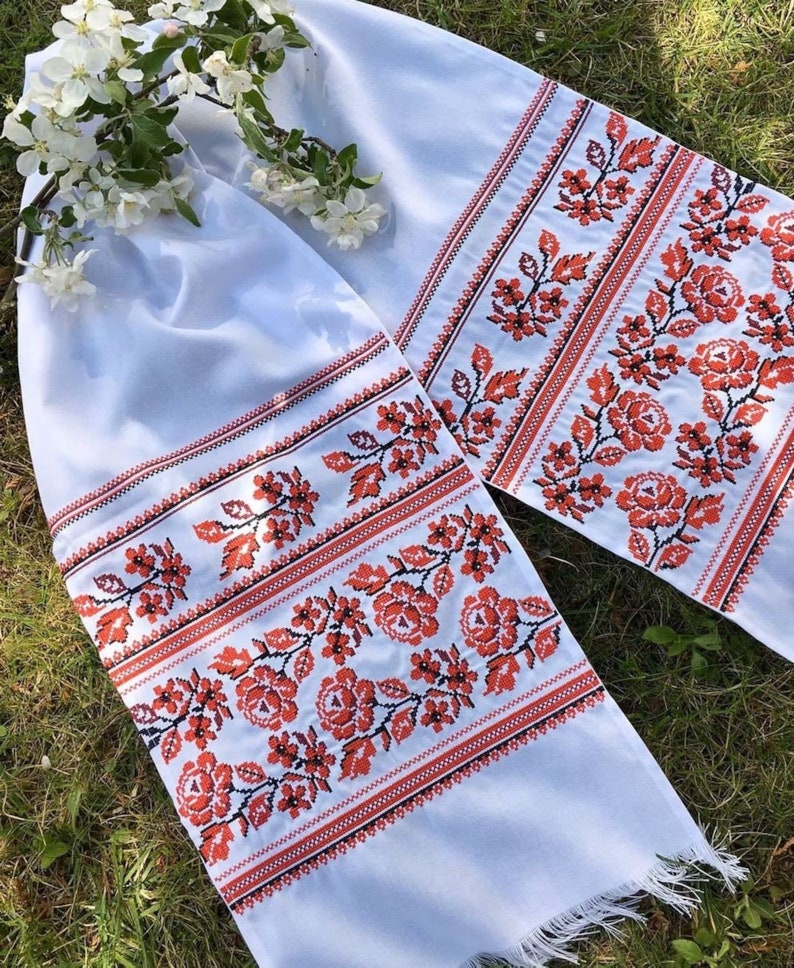 May include: Two white embroidered cloths with red floral patterns and fringed edges. The floral design includes roses and leaves. A sprig of white flowers rests on the cloth, which is displayed on a grassy surface.