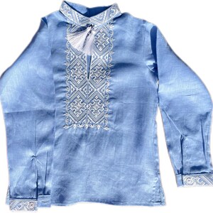 May include: A light blue long-sleeved shirt with intricate white embroidery on the collar and chest. The shirt features a tassel detail and cuffs with matching embroidery. The fabric has a subtle sheen, suggesting a linen or similar material.