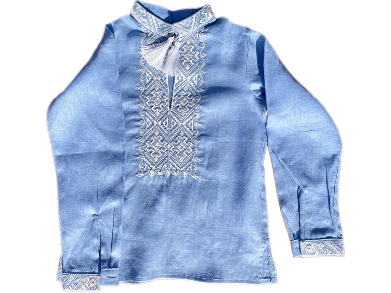 May include: A light blue long-sleeved shirt with intricate white embroidery on the collar and chest. The shirt features a tassel detail and cuffs with matching embroidery. The fabric has a subtle sheen, suggesting a linen or similar material.