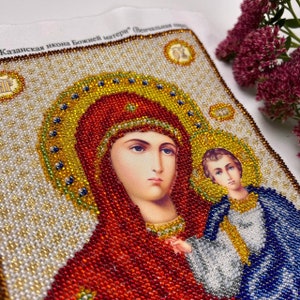 May include: A beaded icon depicting a religious figure with a halo, wearing a red head covering and a gold necklace. The icon features a child figure and is surrounded by gold, white, and blue beads. The text "Казанская икона Божией матери" is visible.