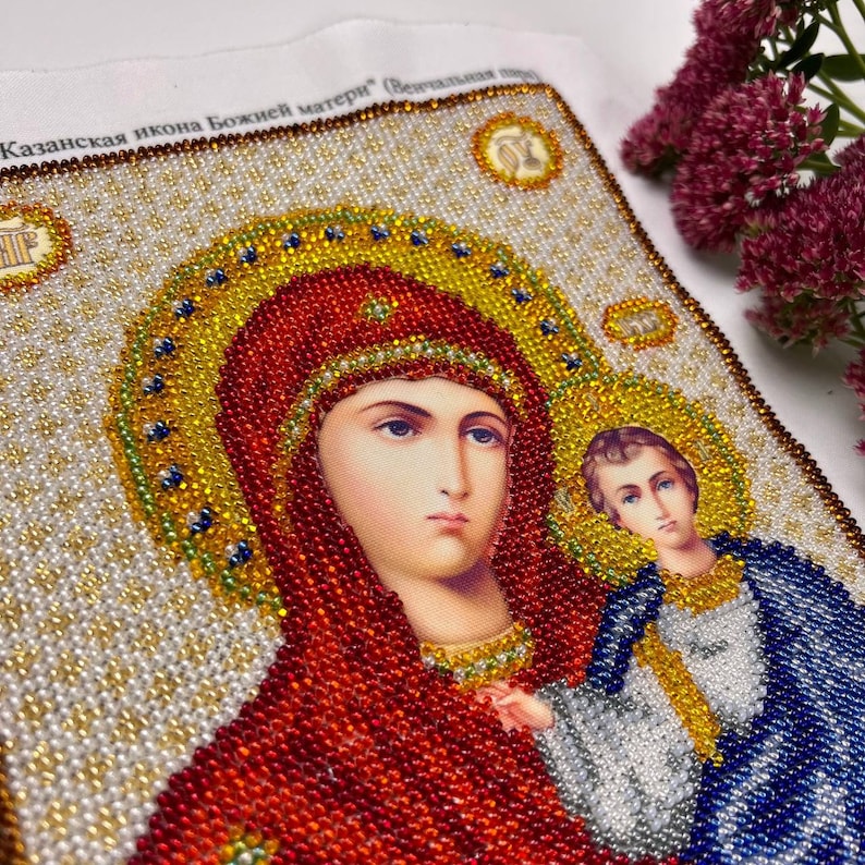May include: A beaded icon depicting a religious figure with a halo, wearing a red head covering and a gold necklace. The icon features a child figure and is surrounded by gold, white, and blue beads. The text "Казанская икона Божией матери" is visible.