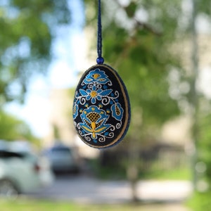K&ouml;nnte beinhalten: Ein blauer und gelber gestickter Osterei-Anh&auml;nger, der an einer blauen Schnur h&auml;ngt. Das Ei ist mit einem Blumenmuster verziert.
