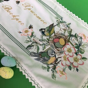以下が含まれることがあります： 白い刺繍のイースターテーブルランナー。花と鳥のデザイン。卵、鳥、花、そして「Christos」と「Voskres」の文字が金色で描かれた巣があります。ランナーは緑色の縁取りとレースのトリムが付いています。