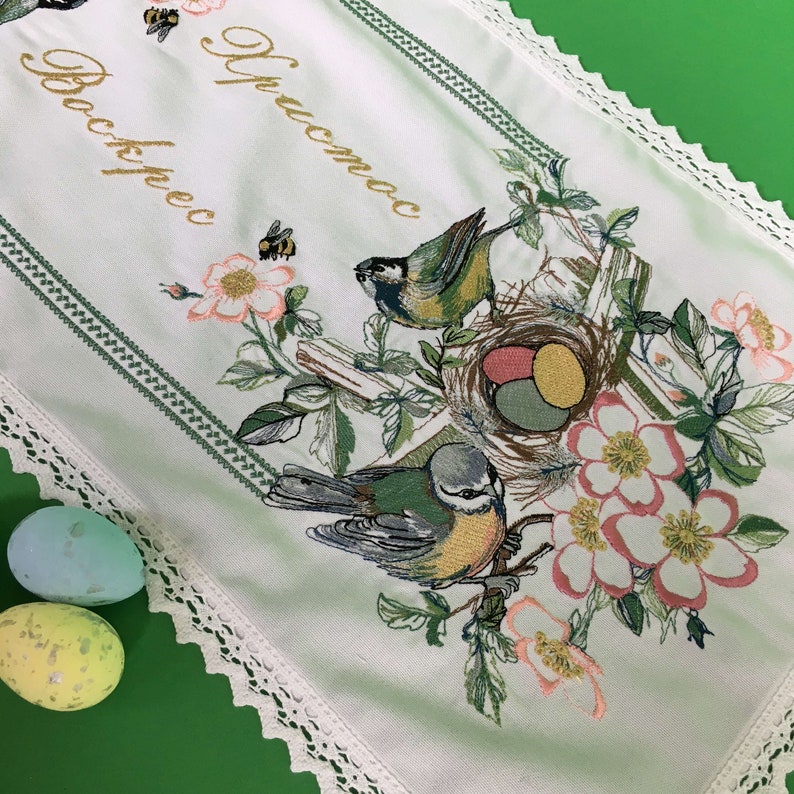 以下が含まれることがあります： 白い刺繍のイースターテーブルランナー。花と鳥のデザイン。卵、鳥、花、そして「Christos」と「Voskres」の文字が金色で描かれた巣があります。ランナーは緑色の縁取りとレースのトリムが付いています。