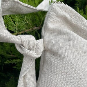 May include: A close-up of a beige canvas tote bag with a zipper closure and a knotted strap. The bag is made of a woven fabric and has a natural, unbleached color.
