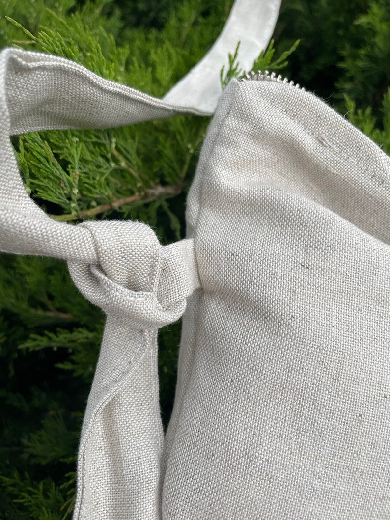 May include: A close-up of a beige canvas tote bag with a zipper closure and a knotted strap. The bag is made of a woven fabric and has a natural, unbleached color.