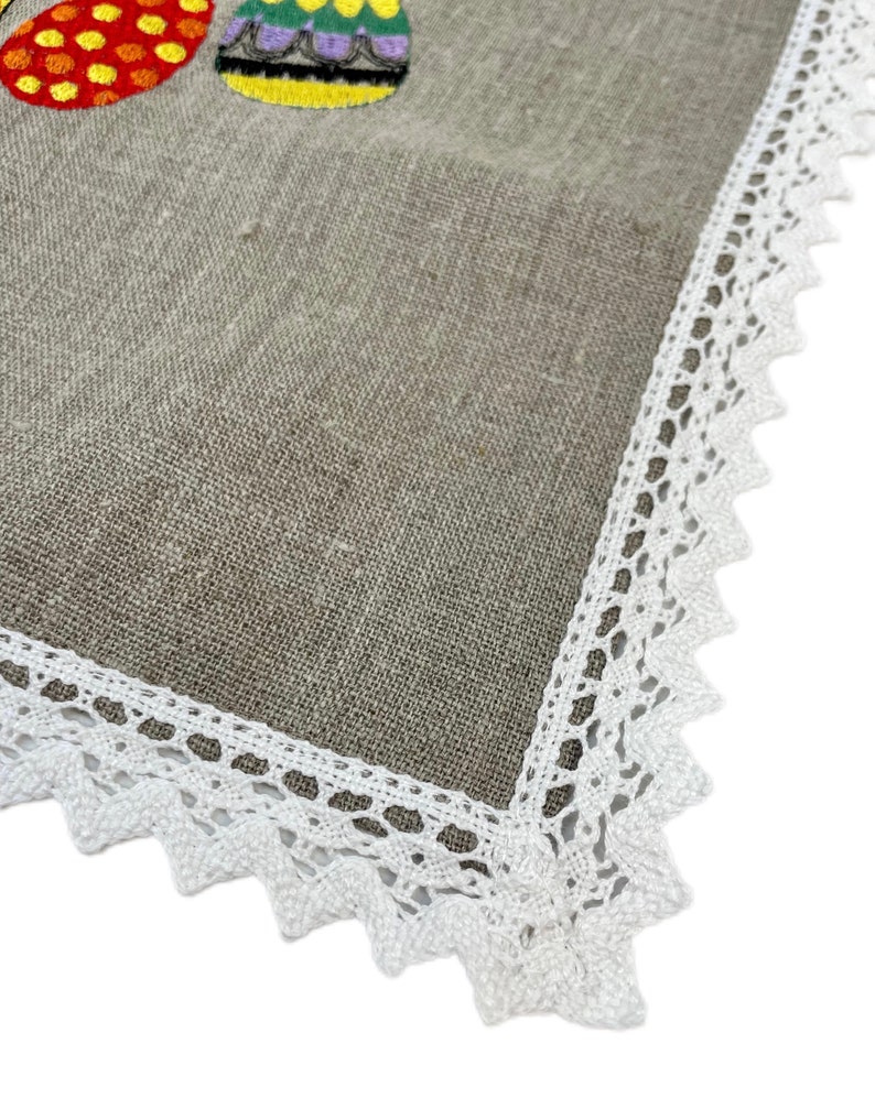 May include: A beige linen tablecloth with a white lace border. The tablecloth has embroidered colorful Easter eggs.