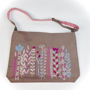 K&ouml;nnte beinhalten: Eine braune Canvas-Tragetasche mit einem rosafarbenen Riemen und einem gestickten Blumendesign. Die Blumen sind in den Farben Rosa, Blau und Wei&szlig; gehalten.