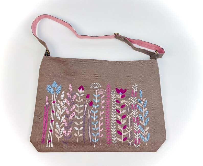 K&ouml;nnte beinhalten: Eine braune Canvas-Tragetasche mit einem rosafarbenen Riemen und einem gestickten Blumendesign. Die Blumen sind in den Farben Rosa, Blau und Wei&szlig; gehalten.