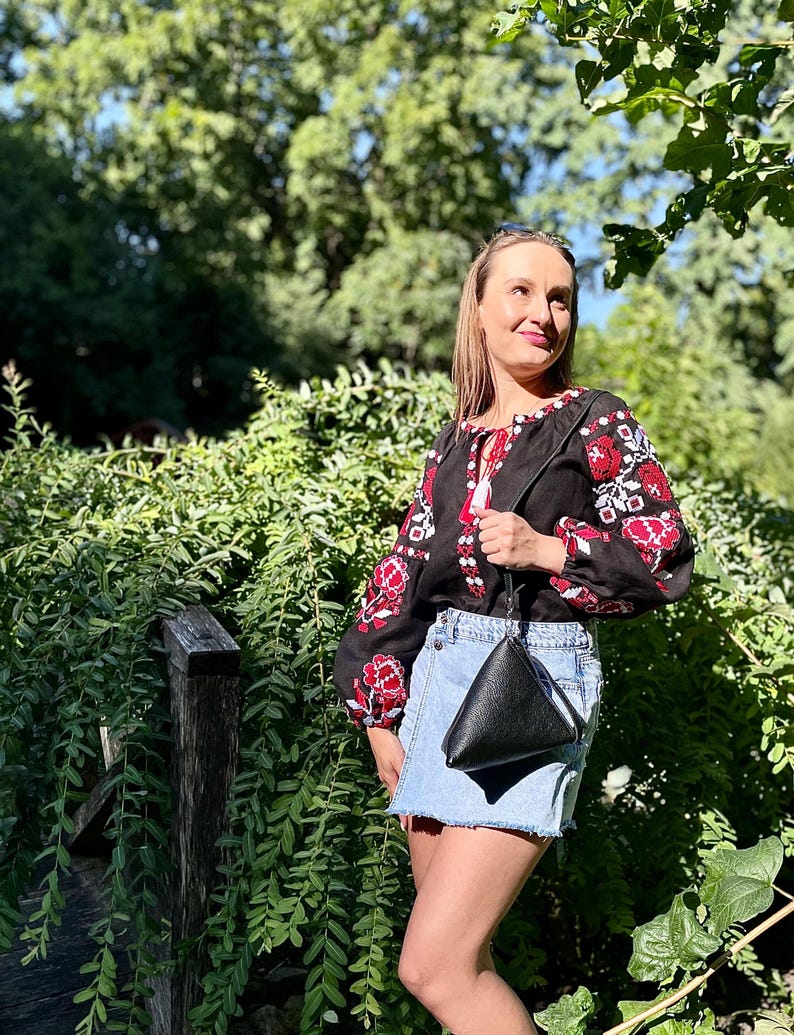 May include: A woman wearing a black embroidered blouse with red and white floral patterns, a denim skirt, and a black triangular handbag.