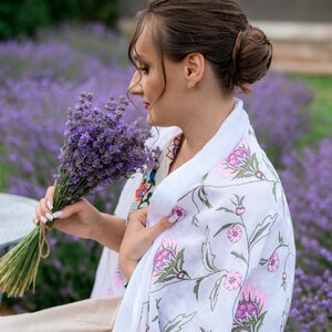 May include: A woman holding a bunch of lavender in a field of purple flowers. She is wearing a white shawl with pink and purple floral embroidery. The shawl is draped over her shoulders.