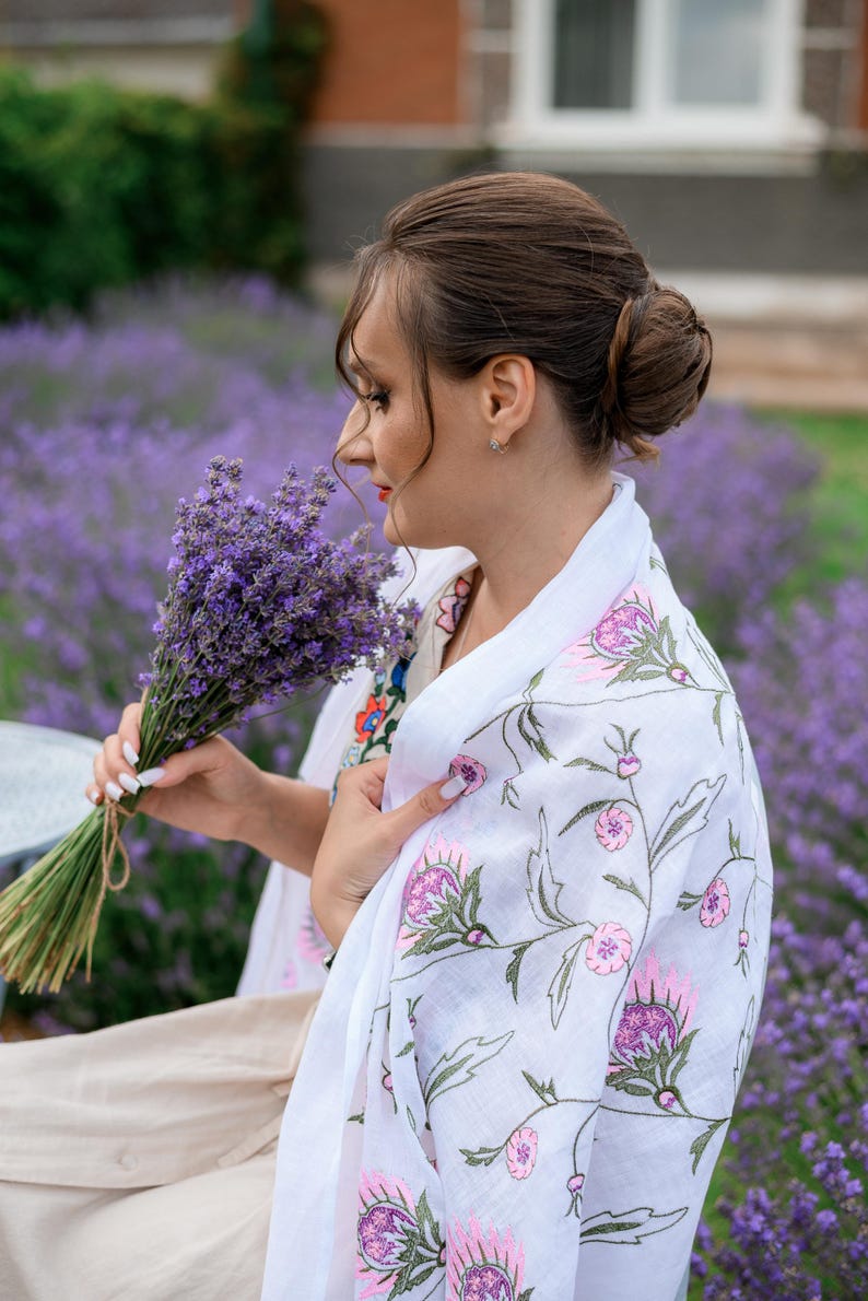 May include: A woman holding a bunch of lavender in a field of purple flowers. She is wearing a white shawl with pink and purple floral embroidery. The shawl is draped over her shoulders.