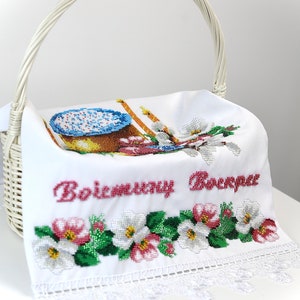 K&ouml;nnte beinhalten: Wei&szlig;er Weidenkorb mit Henkel, der ein wei&szlig;es, besticktes Tuch h&auml;lt. Das Tuch zeigt ein Kreuzstichmuster aus Kuchen, Kerzen und Blumen. Der Text "Воістину Воскрес" ist rot gestickt.