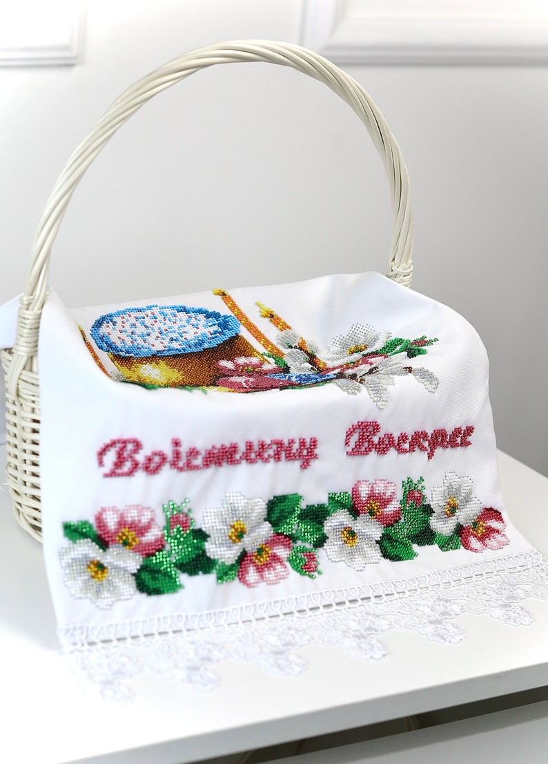 K&ouml;nnte beinhalten: Wei&szlig;er Weidenkorb mit Henkel, der ein wei&szlig;es, besticktes Tuch h&auml;lt. Das Tuch zeigt ein Kreuzstichmuster aus Kuchen, Kerzen und Blumen. Der Text "Воістину Воскрес" ist rot gestickt.