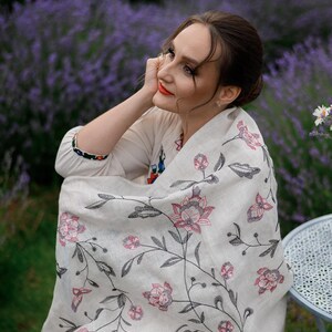 May include: A woman wearing a white shawl with a floral embroidery pattern in pink and black. The shawl is draped over her shoulders and she is sitting in a field of lavender.
