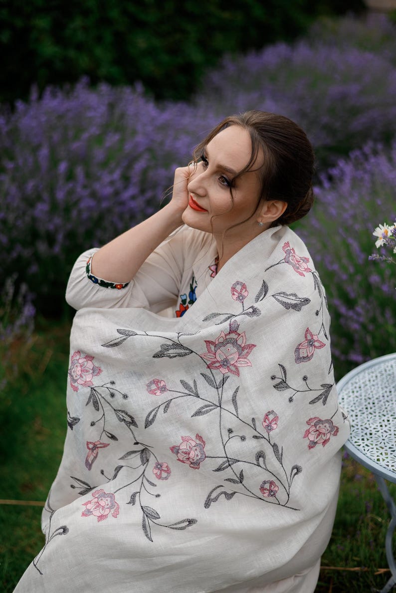 May include: A woman wearing a white shawl with a floral embroidery pattern in pink and black. The shawl is draped over her shoulders and she is sitting in a field of lavender.