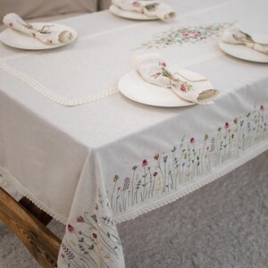 May include: A white linen tablecloth with a delicate lace trim and embroidered floral design. The tablecloth is set with white plates and napkins.
