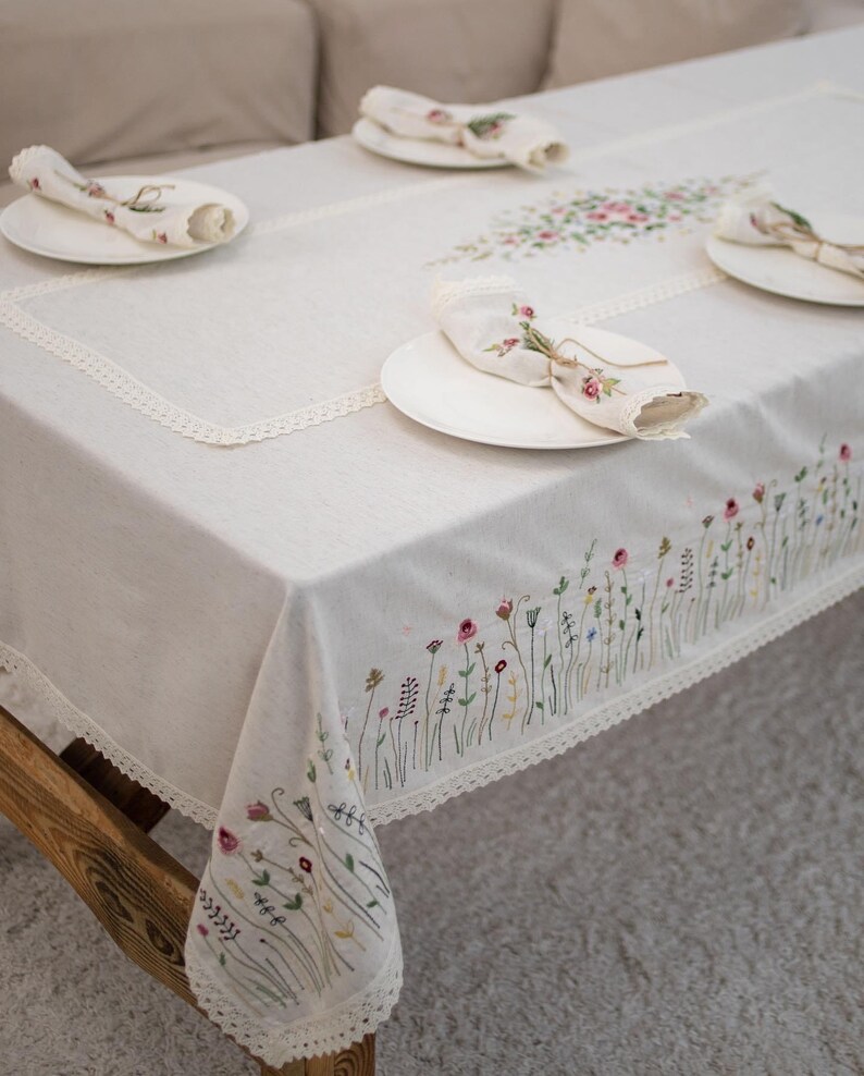 May include: A white linen tablecloth with a delicate lace trim and embroidered floral design. The tablecloth is set with white plates and napkins.