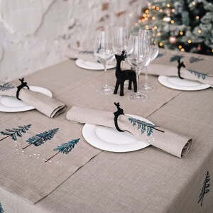 Op de afbeelding: Een beige linnen tafelkleed met een blauw en groen geborduurd dennenboomontwerp. De tafel is gedekt met witte borden en servetten met zwarte rendier servetringen.