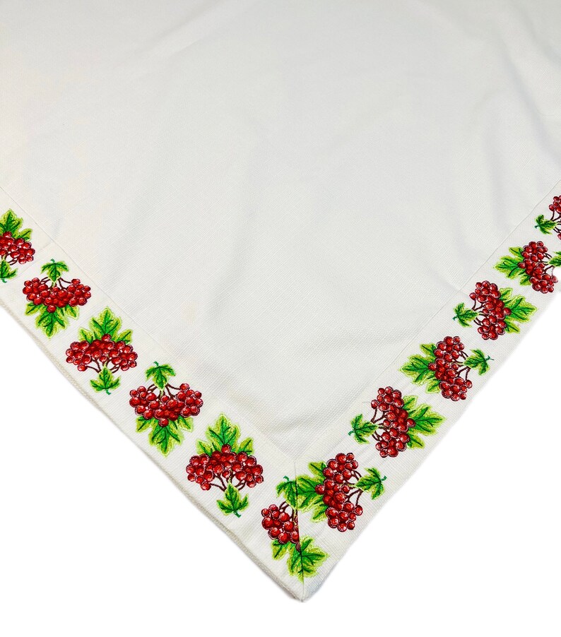 May include: White tablecloth with a detailed embroidered border featuring clusters of red berries and green leaves. The embroidery is on the edge of the cloth, creating a decorative frame.