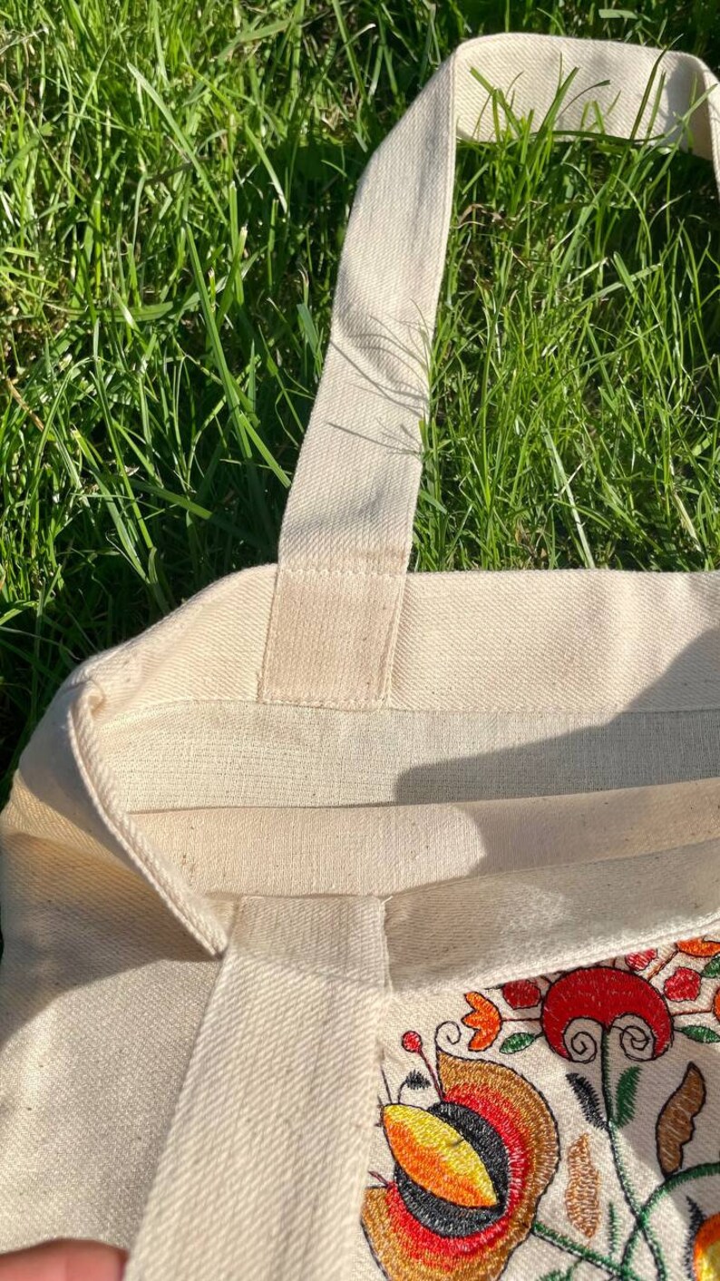 May include: A natural canvas tote bag with a floral embroidery design in shades of red, orange, yellow, and green. The bag has a single strap and is made of a sturdy fabric.