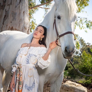 May include: A woman in a white off-the-shoulder dress with a floral print stands next to a white horse. She is holding the horse's reins and looking at the camera.