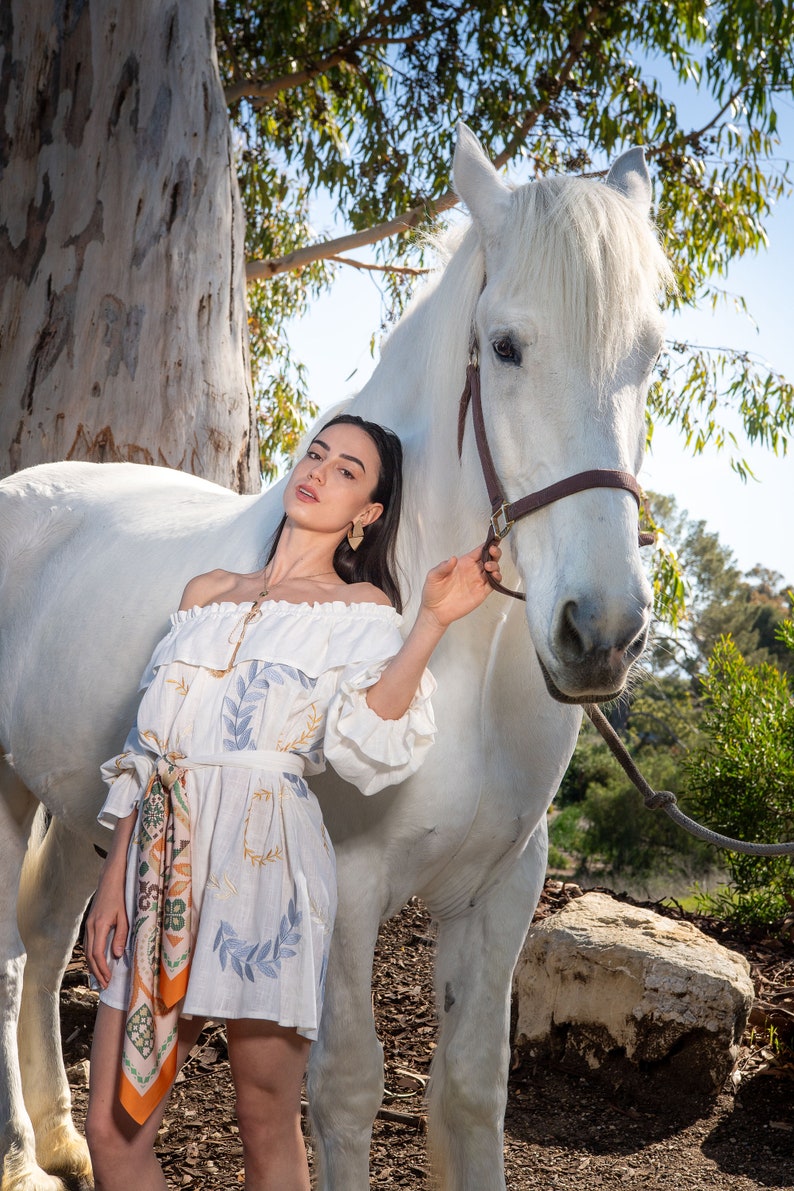 May include: A woman in a white off-the-shoulder dress with a floral print stands next to a white horse. She is holding the horse's reins and looking at the camera.