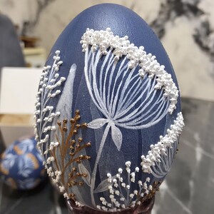 Puede incluir: Un huevo decorativo azul marino con dise&ntilde;os florales blancos y dorados. El huevo presenta una gran flor blanca con p&eacute;talos detallados y peque&ntilde;os acentos florales blancos. Los tallos y hojas dorados a&ntilde;aden contraste.