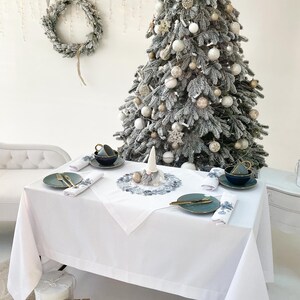 K&ouml;nnte beinhalten: Eine festliche Weihnachtsdekoration mit wei&szlig;er Tischdecke, teal-farbenen Tellern und goldenem Besteck. Im Hintergrund ein geschm&uuml;ckter Weihnachtsbaum, sowie ein Kranz und Sternschmuck. Geschenkboxen stehen auf dem Boden.