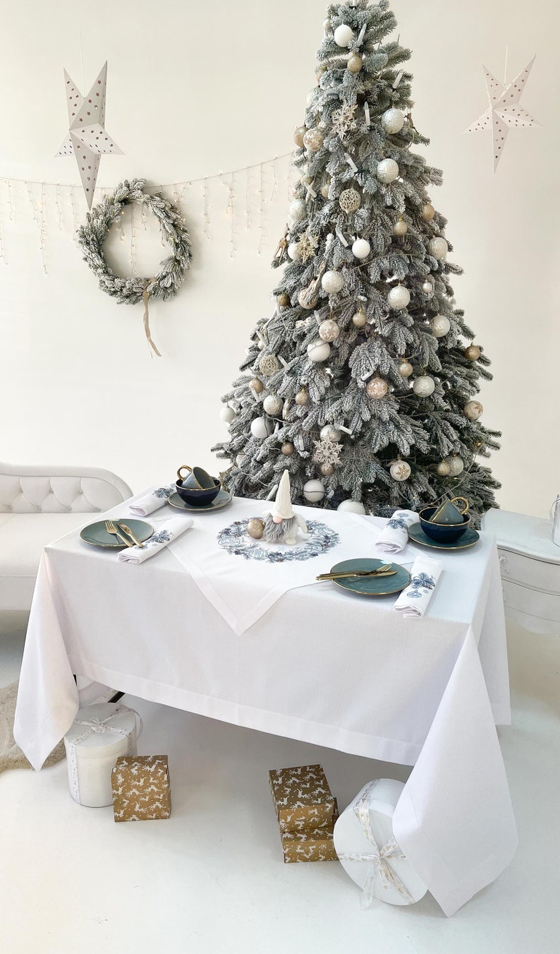 K&ouml;nnte beinhalten: Eine festliche Weihnachtsdekoration mit wei&szlig;er Tischdecke, teal-farbenen Tellern und goldenem Besteck. Im Hintergrund ein geschm&uuml;ckter Weihnachtsbaum, sowie ein Kranz und Sternschmuck. Geschenkboxen stehen auf dem Boden.
