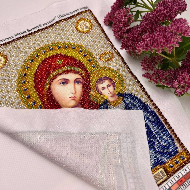 May include: A detailed, beaded icon featuring the Virgin Mary and Child, with halos and ornate detailing. The artwork uses red, gold, blue, and white beads. The text in the image is in a foreign language. The icon is on a white fabric.