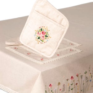 May include: A beige linen pot holder with a floral embroidery design featuring pink roses and blue accents. The pot holder is held up by a hand, showcasing its loop for hanging. A matching tablecloth with embroidered flowers and lace trim is also visible.