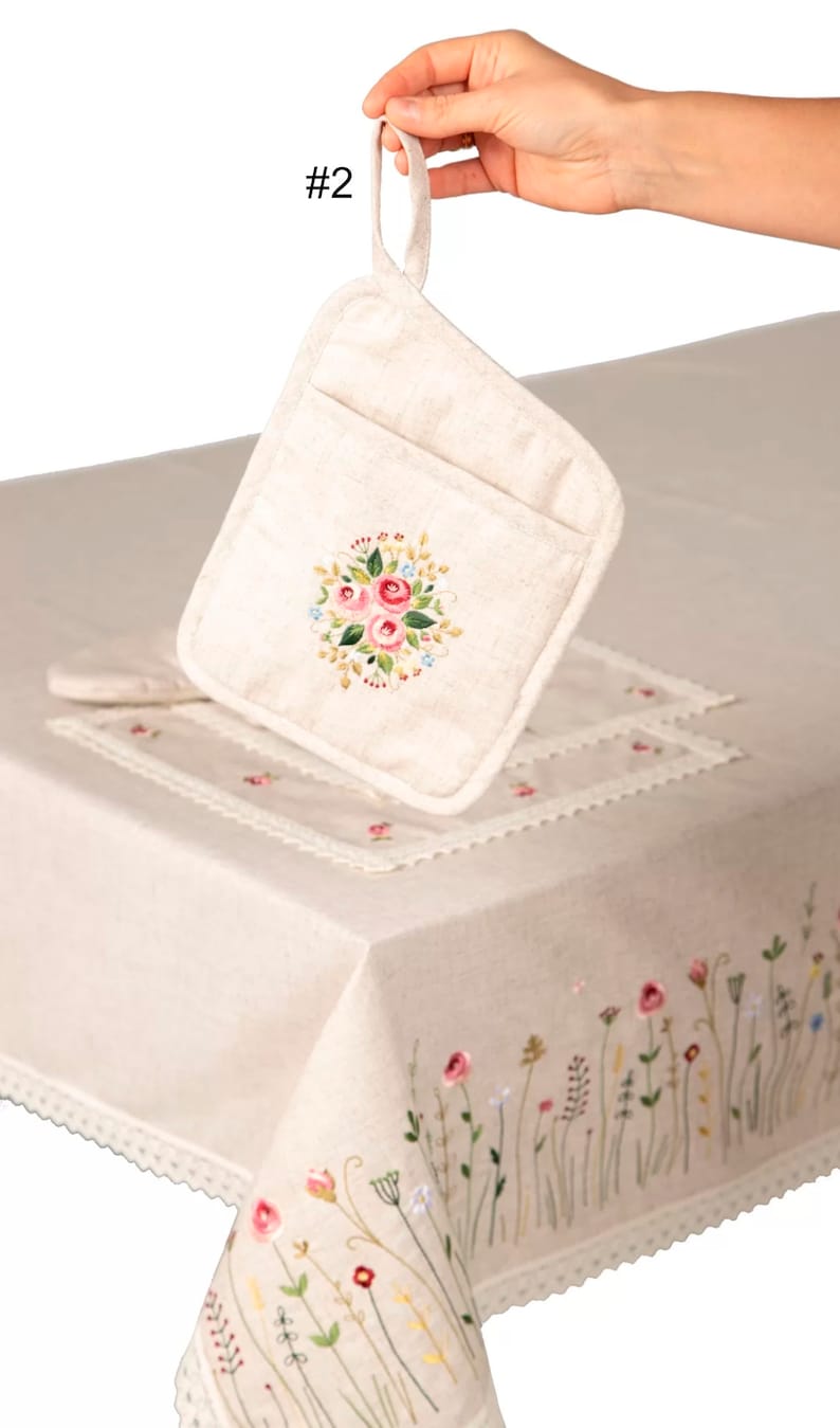 May include: A beige linen pot holder with a floral embroidery design featuring pink roses and blue accents. The pot holder is held up by a hand, showcasing its loop for hanging. A matching tablecloth with embroidered flowers and lace trim is also visible.