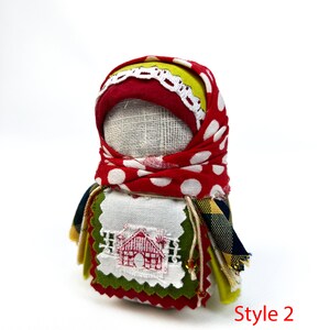 Op de afbeelding: Een handgemaakte stoffen pop met een rood-wit geblokt hoofddoekje, een groen-gele geruite rok en een wit geborduurd plekje met een rood huisontwerp. De pop is gemarkeerd met "Style 2".