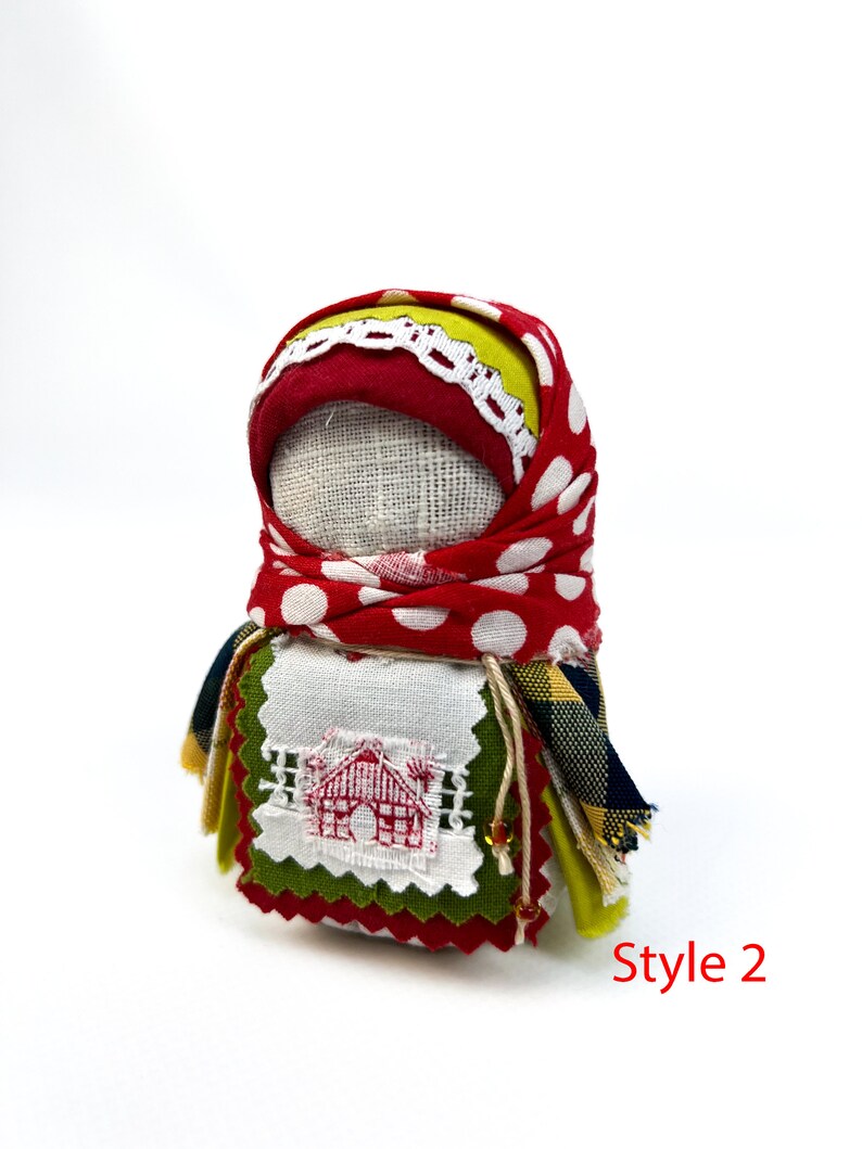 Op de afbeelding: Een handgemaakte stoffen pop met een rood-wit geblokt hoofddoekje, een groen-gele geruite rok en een wit geborduurd plekje met een rood huisontwerp. De pop is gemarkeerd met "Style 2".