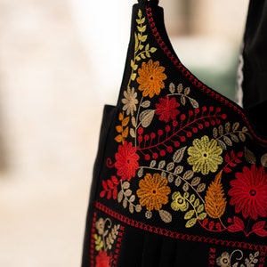 May include: Black shoulder bag with vibrant floral embroidery in red, orange, and yellow. The bag features a detailed design of flowers and leaves, with a contrasting red stitched border. The strap is also black.