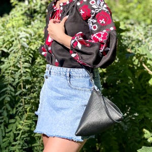 May include: A woman wearing a black embroidered blouse with red and white floral patterns and a light blue denim skirt. She is carrying a black leather triangular shaped purse.
