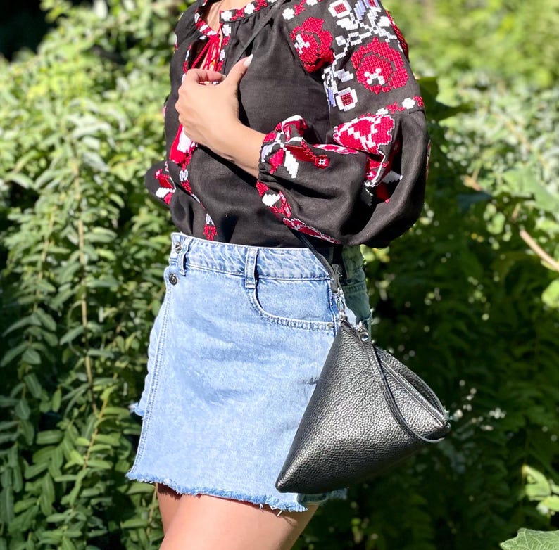 May include: A woman wearing a black embroidered blouse with red and white floral patterns and a light blue denim skirt. She is carrying a black leather triangular shaped purse.