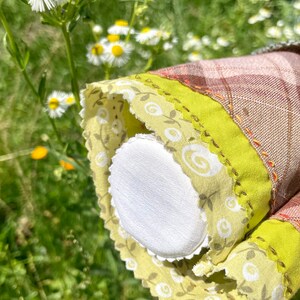 Puede incluir: Primer plano de un c&iacute;rculo de tela blanca con un ribete floral verde y blanco. El ribete tiene un borde festoneado y est&aacute; cosido sobre una tela de cuadros marr&oacute;n y blanco.