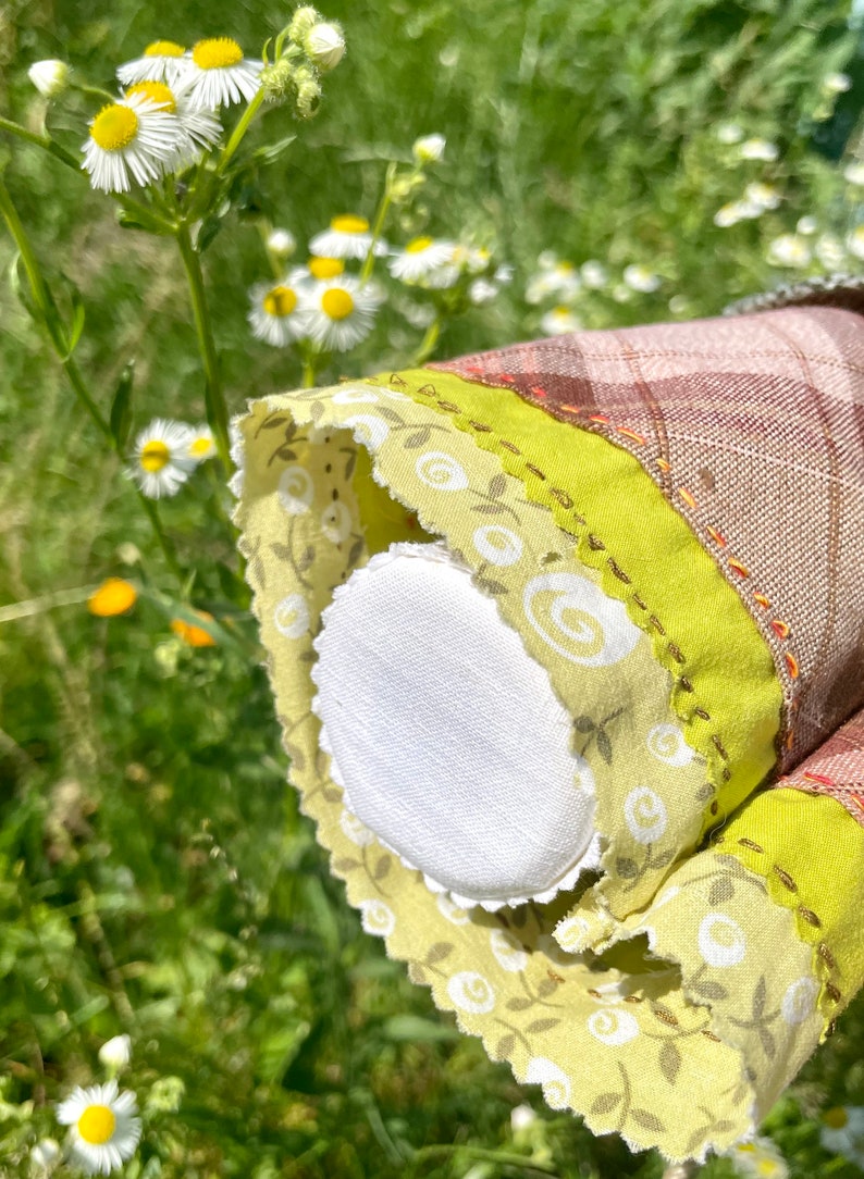 Puede incluir: Primer plano de un c&iacute;rculo de tela blanca con un ribete floral verde y blanco. El ribete tiene un borde festoneado y est&aacute; cosido sobre una tela de cuadros marr&oacute;n y blanco.