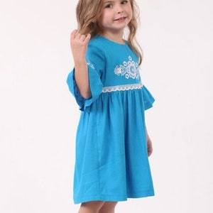 K&ouml;nnte beinhalten: Ein junges M&auml;dchen tr&auml;gt ein blaues Kleid mit wei&szlig;en Stickereien und einem wei&szlig;en Spitzenbesatz. Das Kleid hat einen R&uuml;schen-Saum und 3/4-&Auml;rmel.